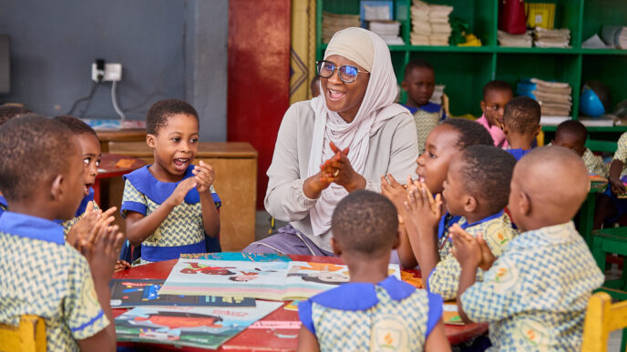 Children in green and blue uniform, sat round a table looking up a teacher wearing a hijab and smiling at them.