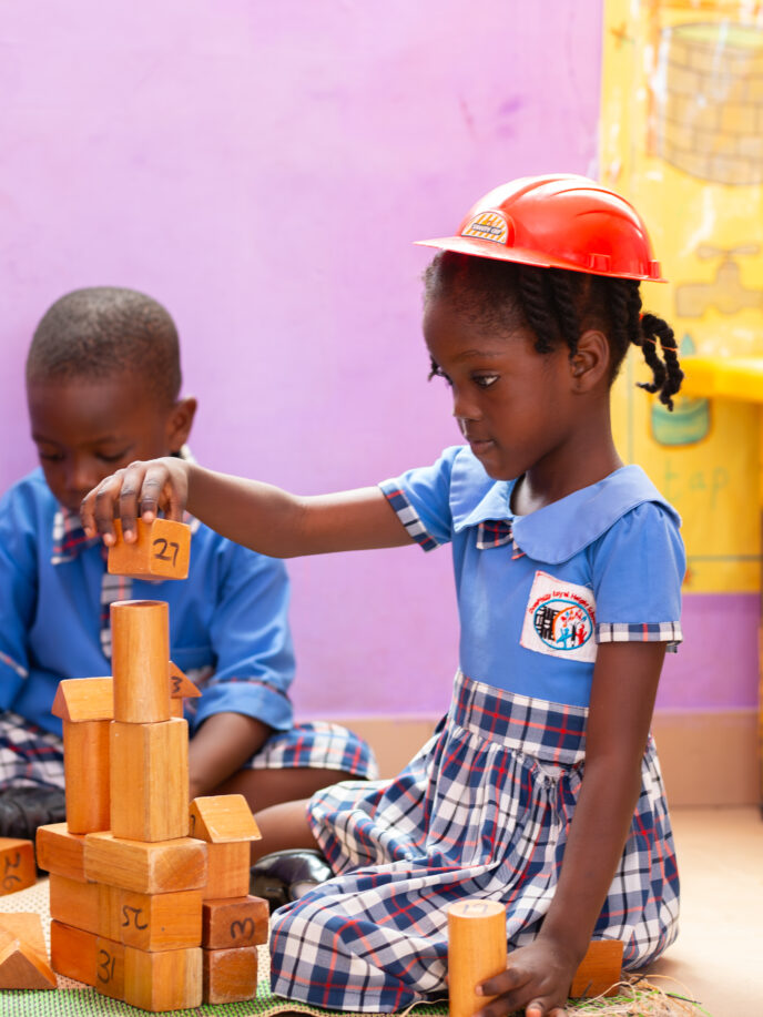 A boy and a girl playing with construction blocks dressed in blue and gingham uniforms the girls has a red helmet on her head